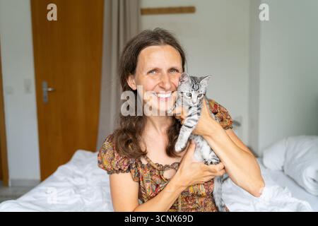Eine Frau hält ein verspieltes graues Kätzchen in den Armen und lächelt warm, während sie auf einem Bett sitzt. Die gemütliche Wohnatmosphäre verleiht Ihrem Zuhause ein Gefühl von Komfort und Freude Stockfoto