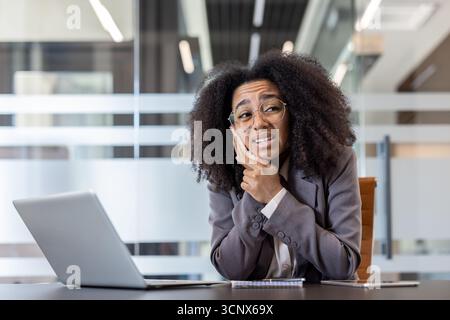 Eine traurige Afroamerikanerin sitzt an einem Schreibtisch im Büro und hält ihre Wange mit der Hand und spürt schwere Zahnschmerzen. Stockfoto