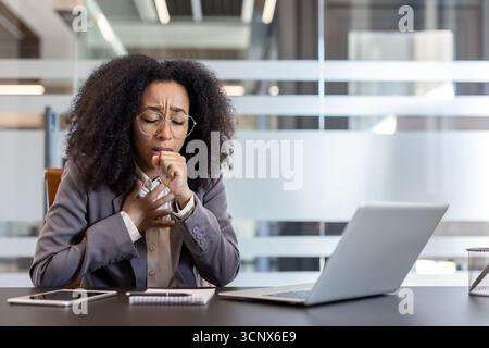 Junge afroamerikanische Geschäftsfrau, die an Asthmaanfällen leidet und hinter ihrem Arbeitsplatz im Büro hustet. Stockfoto