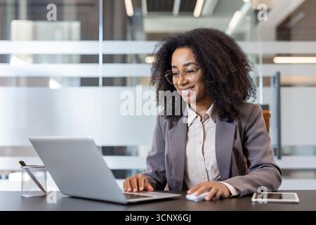 Erfolgreiche und lächelnde junge afroamerikanische Frau, die im Büro an einem Laptop arbeitet und ein Online-Meeting abhält. Stockfoto