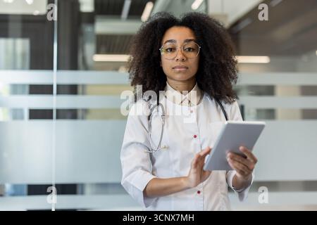 Porträt einer ernsthaften jungen afroamerikanischen Ärztin in Uniform und Brille, die in der Klinik steht, Tablette hält und in die Kamera blickt. Stockfoto
