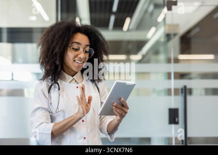 Eine junge afroamerikanische Ärztin steht in einem weißen Mantel in der Klinik und spricht bei einem Videoanruf mit der Kamera des Tablets, das sie in den Händen hält. Stockfoto