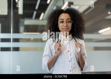 Porträt einer ernsthaften jungen afroamerikanischen Ärztin, Wissenschaftlerin, Apotheker, die in Uniform in der Klinik steht, das Stethoskop in den Händen hält und selbstbewusst in die Kamera blickt. Stockfoto