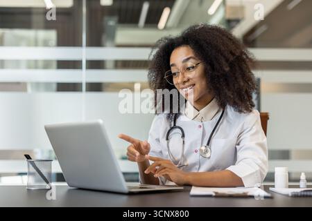 Eine junge afroamerikanische Ärztin sitzt an einem Schreibtisch in einem Klinikbüro und spricht auf einem Laptop über ein Online-Videogespräch. Stockfoto
