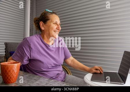 Eine ältere Frau sitzt bequem zu Hause und lächelt, während sie an ihrem Laptop arbeitet. Sie trägt ein lila Hemd und scheint zufrieden zu sein. Sie verkörpert einen entspannten Modus Stockfoto