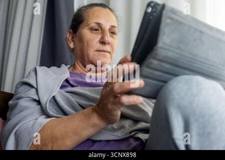Seniorin sitzt bequem zu Hause und interagiert mit einem digitalen Tablet. Die Szene reflektiert einen Moment der Freizeit und moderner Konnektivität in einem entspannten Stockfoto