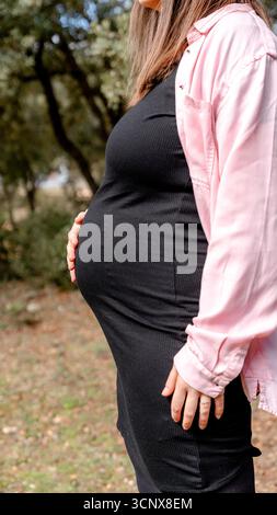 Abgeschnittene, nicht erkennbare Seitenansicht einer schwangeren Mutter in der 35. Schwangerschaftswoche, die sanft ihren Bauch hält, während sie in einer ruhigen Landschaft steht Stockfoto