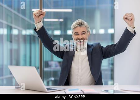 Ein Geschäftsmann, der mit Fäusten in einem modernen Büro hochgezogenen, feiert Karriereerfolg und einen großen Sieg. Selbstbewusster, freudiger Manager zeigt Triumph und Motivation an seinem Schreibtisch Stockfoto