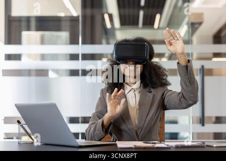 Junge afroamerikanische Frau, die im Büro am Schreibtisch sitzt, in einem Anzug, mit virtueller Brille und mit den Händen Gesten trägt. Stockfoto