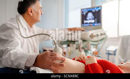 Ein Gynäkologe führt eine Ultraschalluntersuchung bei einer schwangeren Frau durch und beobachtet die fetale Entwicklung am Monitor. Die Einstellung ist klinisch, wobei die pränatale Einstellung hervorgehoben wird Stockfoto