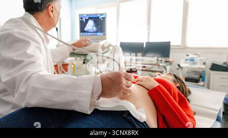 Ein Gynäkologe führt eine Ultraschalluntersuchung an einer schwangeren Frau in einer Arztpraxis durch. Die werdende Mutter liegt auf einem Untersuchungstisch und beobachtet Stockfoto