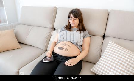 Eine schwangere Frau im dritten Trimenon sitzt bequem auf einem Sofa und zeigt eine verspielte Ladegrafik auf ihrem Bauch. Sie hat einen Ultraschall-ph-wert Stockfoto