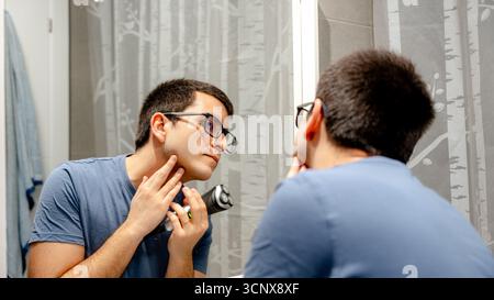 Mann, der vor einem Badezimmerspiegel steht und sich aufmerksam seinen Bart mit einem Elektrorasierer rasiert. Er trägt eine Brille und ein blaues Hemd, um einen Preci zu gewährleisten Stockfoto