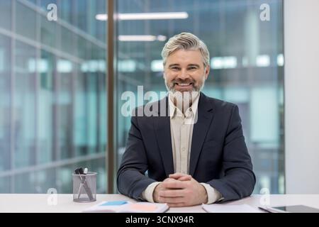 Ein reifer, bärtiger Geschäftsmann sitzt an seinem Schreibtisch, lächelt selbstbewusst und blickt in die Kamera. Er vermittelt Professionalität, Erfolg und Vertrauen in eine moderne Unternehmensumgebung Stockfoto