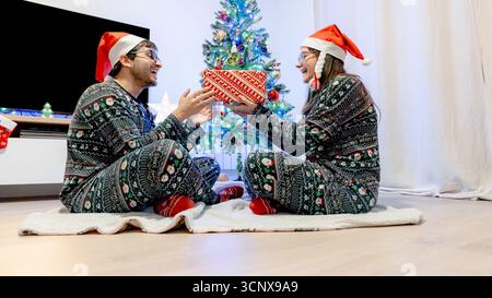 Ein fröhliches Paar im passenden Weihnachts-Pyjama tauscht ein wunderschön gewickeltes Geschenk vor einem glitzernden Baum aus. Der gemütliche Feiertagsgeist fängt mich ein Stockfoto