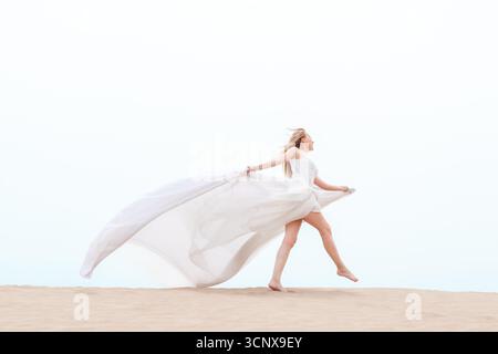 Eine elegante junge Frau in einem fließenden weißen Kleid tanzt anmutig am Strand und verkörpert Freiheit und Eleganz. Das Kleid weht im Wind, Enhanci Stockfoto