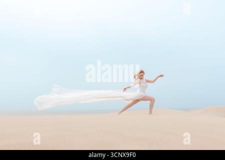 Elegante junge Frau tanzt anmutig an einem Sandstrand, ihr fließendes weißes Kleid weht im Wind. Die Szene fängt Schönheit, Bewegung und Freiheit ein Stockfoto