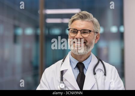 Lächelnder reifer Arzt mit weißem Labormantel und Stethoskop, der selbstbewusst in einer modernen Gesundheitsumgebung wegblickt und Weisheit und professionelle Pflege repräsentiert Stockfoto
