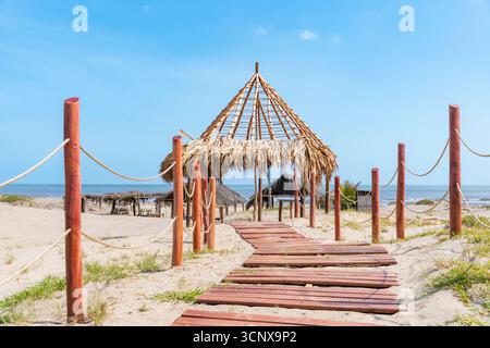 Ein malerischer Blick auf den Strand Punta Astilleros in Kolumbien mit einem Holzsteg, der zu einem strohgedeckten Hüttenpavillon am Meer unter einem klaren blauen Himmel führt. Stockfoto
