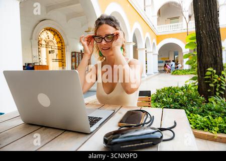Eine junge Frau arbeitet ferngesteuert an einem Laptop in einem hellen Innenhof. Sie passt ihre Brille an und fokussiert dabei intensiv auf den Bildschirm, um das digitale n zu berücksichtigen Stockfoto