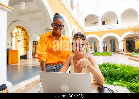 Eine multirassische Gruppe junger digitaler Nomadenfrauen beschäftigt sich mit Arbeit und Entspannung. Eine Kaukasierin und eine Schwarze arbeiten an einem Laptop in einem sunn zusammen Stockfoto