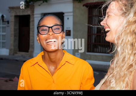 Ein freudiger Moment zwischen zwei jungen multiethnischen Frauen, einer schwarzen und einer kaukasischen Frau, die auf einer lebhaften Straße lachen und ihren Freund umarmen Stockfoto