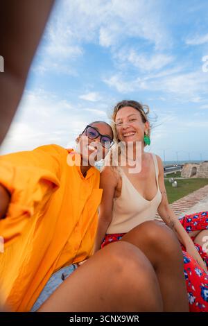 Zwei junge multiethnische Frauen, eine schwarze und eine kaukasierin, machen glücklich ein Selfie und halten einen freudigen Moment vor einem wunderschönen blauen Himmel in Cartagena d fest Stockfoto