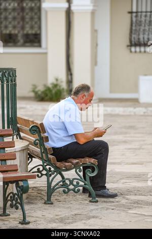 Ein älterer Mann sitzt allein auf einer Bank im Stadtzentrum mit einem Smartphone oder Mobiltelefon. Ein älterer oder mittelalter Mann, der auf das Telefon schaut. Stockfoto