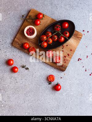 Ein hölzernes Schneidebrett zeigt lebhafte Kirschtomaten, einige ganze und einige halbierte, neben einer kleinen Schüssel mit rosa Pfefferkörnern. Frisch und appetitlich Stockfoto