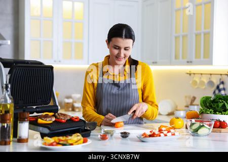 Frau, die Pfeffer schneidet, während sie Fleisch und Gemüse auf einem elektrischen Grill in der Küche kocht Stockfoto