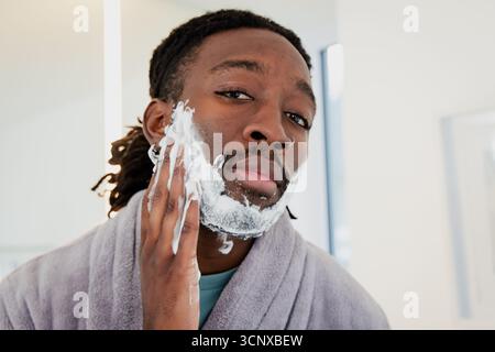 Afroamerikanischer Mann, der Rasierschaum auf das Gesicht im Badezimmer vor dem Spiegel aufträgt und Bademantel trägt Stockfoto