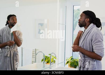 Afroamerikanischer Mann mit Bademantel, trocknendem Gesicht und weißem Handtuch am Badezimmerspiegel, Kopierraum Stockfoto