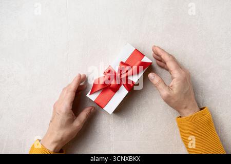 Nahaufnahme der Hände einer Frau in einem gemütlichen gelben Pullover, der ein wunderschön gewickeltes Geschenk mit rotem Band auf hellem Steinhintergrund hält. Perfekt für Chr Stockfoto