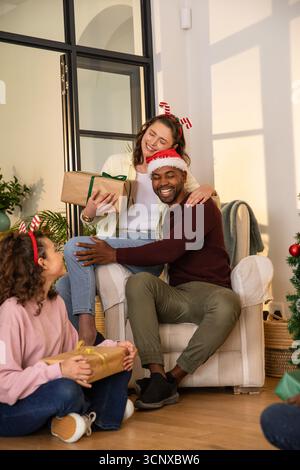 Vielfältige Familiengeschenke in gemütlichem Wohnzimmer mit weihnachtsmütze und Haarbändern Stockfoto