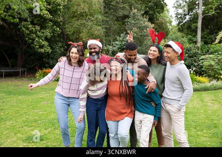Vielfältige, generationsübergreifende Familie, die auf üppigem Rasen im Garten steht und weihnachtsmützen und Geweihe trägt Stockfoto