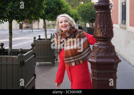 Grauhaarige Frau von 60 Jahren in einem roten Mantel und einem Terrakotta-Schal in der Stadt Stockfoto
