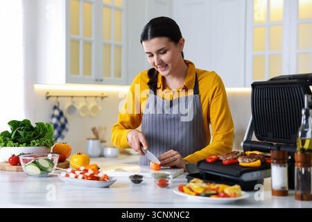 Frau, die Pfeffer schneidet, während sie Fleisch und Gemüse auf einem elektrischen Grill in der Küche kocht Stockfoto