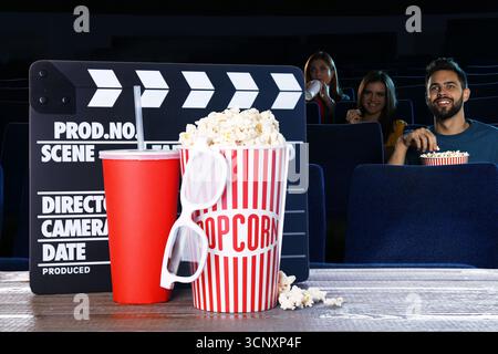 Eimer mit frischem Popcorn, eine Tasse Getränk, 3D-Gläser und eine Klapptafel auf dem Tisch vor Leuten, die sich Filme im dunklen Kino ansehen Stockfoto