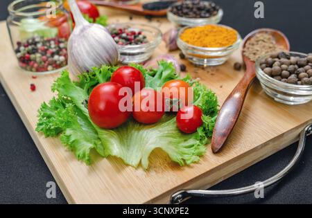 Holzbrett mit Gewürzen und Gemüse. Kirschtomaten, Knoblauch, trockene Gewürze in Schüsseln und Löffeln, an Bord auf schwarzem Hintergrund. Gesundes Essen, Kochen Stockfoto