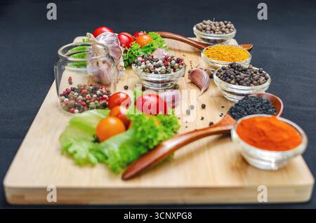 Holzbrett mit Gewürzen und Gemüse. Kirschtomaten, Knoblauch, trockene Gewürze in Schüsseln und Löffeln, an Bord auf schwarzem Hintergrund. Gesundes Essen, Kochen Stockfoto