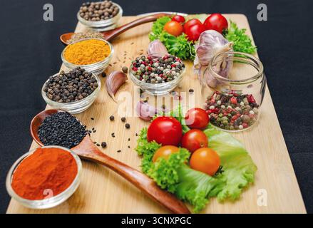Holzbrett mit Gewürzen und Gemüse. Kirschtomaten, Knoblauch, trockene Gewürze in Schüsseln und Löffeln, an Bord auf schwarzem Hintergrund. Gesundes Essen, Kochen Stockfoto