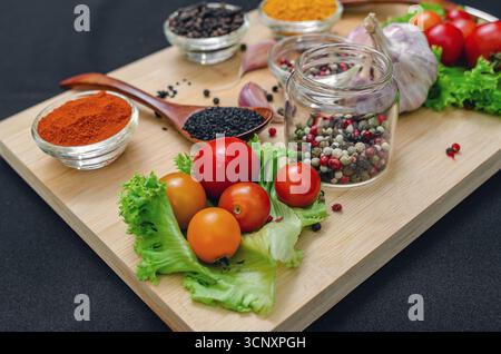 Holzbrett mit Gewürzen und Gemüse. Kirschtomaten, Knoblauch, trockene Gewürze in Schüsseln und Löffeln, an Bord auf schwarzem Hintergrund. Gesundes Essen, Kochen Stockfoto