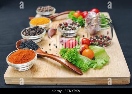 Holzbrett mit Gewürzen und Gemüse. Kirschtomaten, Knoblauch, trockene Gewürze in Schüsseln und Löffeln, an Bord auf schwarzem Hintergrund. Gesundes Essen, Kochen Stockfoto