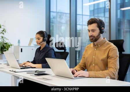 Zwei verschiedene Callcenter-Mitarbeiter, die Headsets tragen und Laptops verwenden, während sie an Schreibtischen in einem hellen, modernen Großraumbüro arbeiten, bieten effizientes Telemarketing und technischen Online-Support Stockfoto