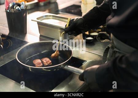 Kaukasier mittleren Alters, der schwarze Handschuhe trägt und Fleischstücke in der Pfanne auf dem Herd in der professionellen Küche anbrennt Stockfoto