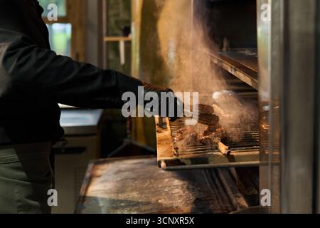 Mann mit mittlerem Alter, der Fleisch auf offener Flamme mit einer Zange grillt, schwarze Handschuhe trägt, am kommerziellen Küchengrill steht, Rauch von der Kochfläche aufsteigt, konzentriert sich auf die Zubereitung Stockfoto