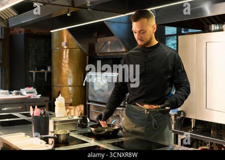 Kaukasischer junger Erwachsener, der Steak in der professionellen Küche kocht, den Teller in einer Hand hält und das Fleisch mit der anderen Hand in die Pfanne legt, konzentriert sich auf die kulinarische Zubereitung Stockfoto