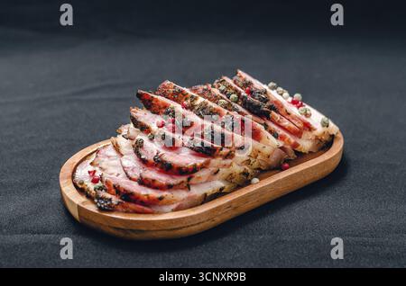 Duftende Scheiben von gebackenem Schweinebauch auf einem Holzbrett, bestreut mit farbigen Pfefferkörnern. Schwarzer Hintergrund. Fleischzubereitungskonzept. Stockfoto