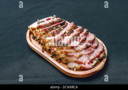Duftende Scheiben von gebackenem Schweinebauch auf einem Holzbrett, bestreut mit farbigen Pfefferkörnern. Schwarzer Hintergrund. Fleischzubereitungskonzept. Stockfoto