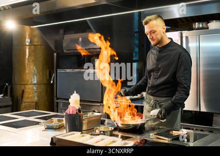 Kaukasischer junger Erwachsener, der in der professionellen Küche kocht, am Herd steht und Pfanne mit großer offener Flamme hält, konzentriert auf die Zubereitung von Lebensmitteln, Edelstahlgeräte sichtbar Stockfoto